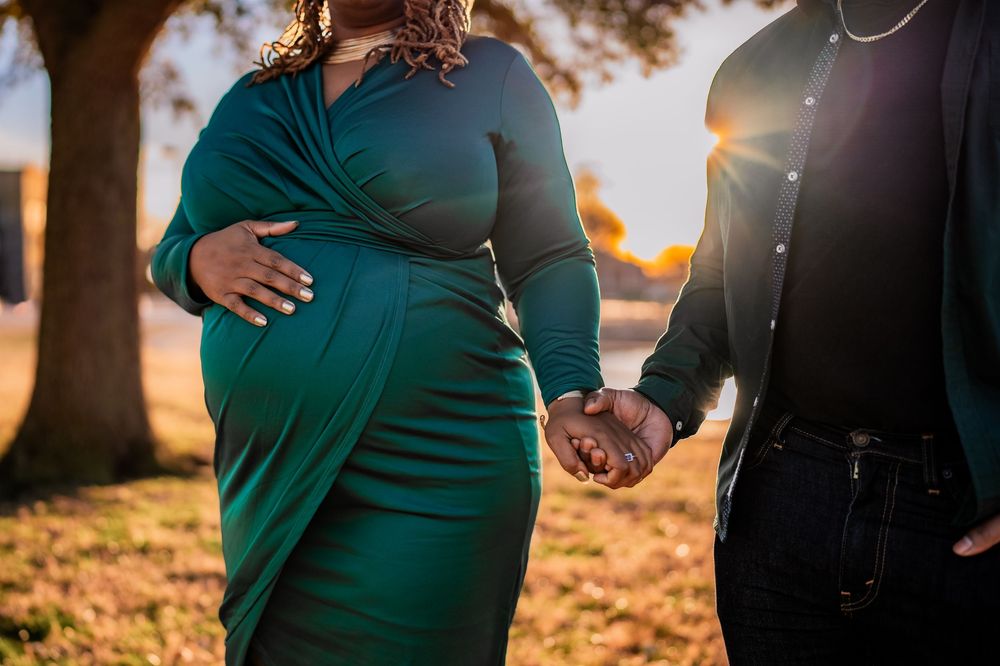 Pregnant woman in green dress holds partner's hand outdoors at golden hour sunset.