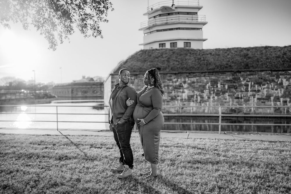 Black and white photo of a pregnant couple posing outdoors near a river lock structure at sunset.