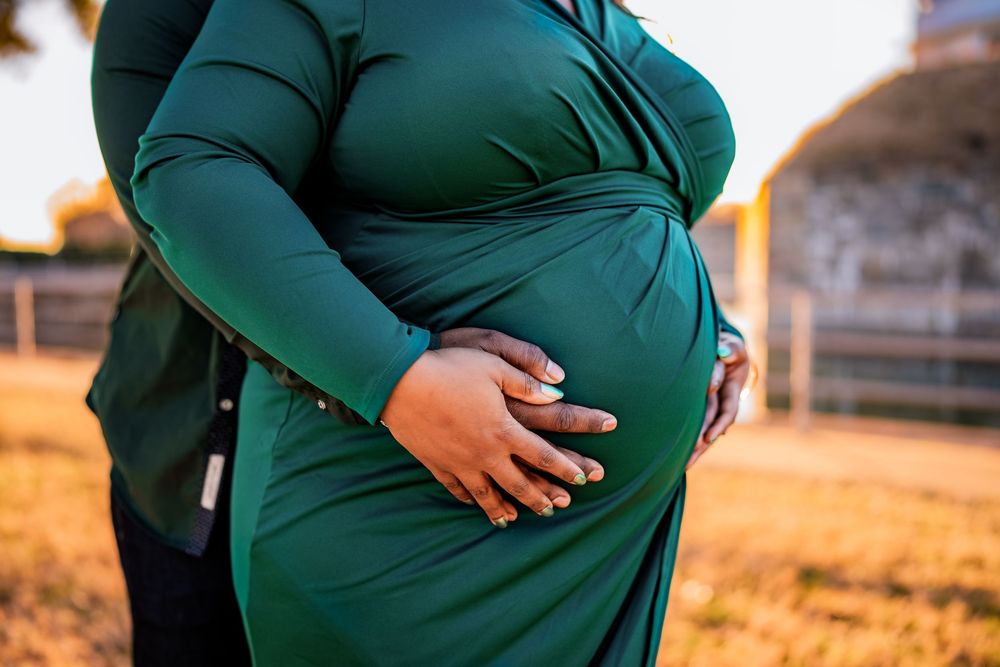 Pregnant woman in green dress cradling baby bump outdoors in golden hour light.