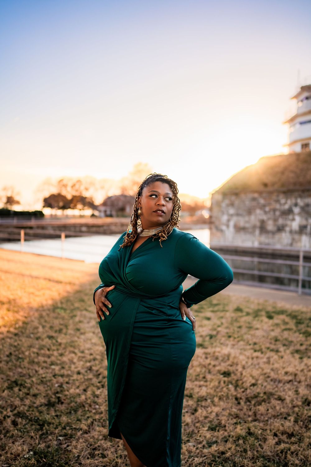 Pregnant Black woman in teal dress poses at sunset near waterway.