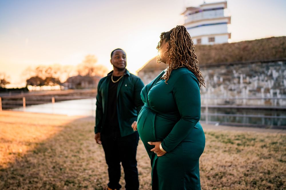 Expecting couple in matching teal outfits standing together at sunset near a historic waterway.