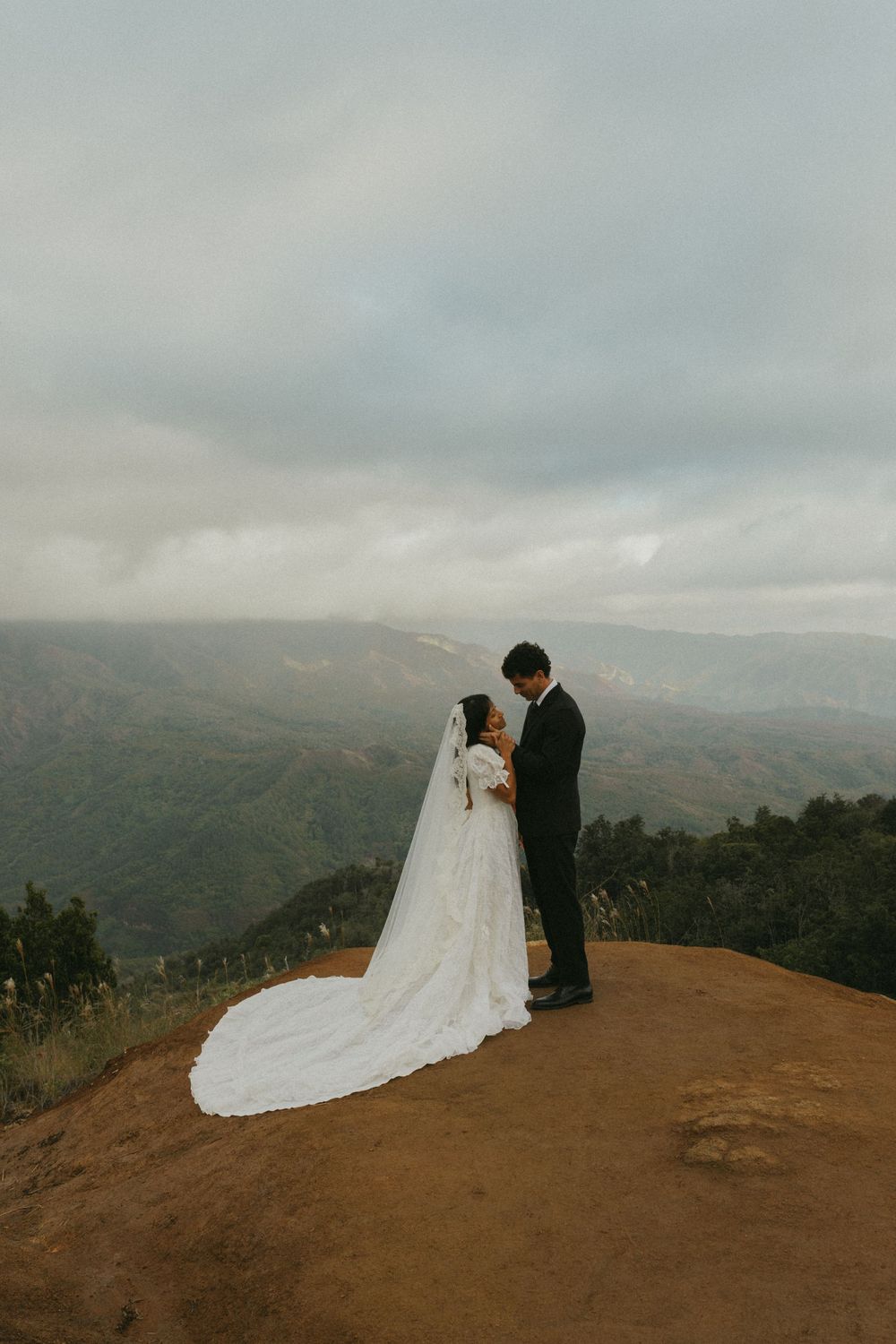 A couple stands together on a scenic mountain overlook with a dramatic cloudy sky and sweeping valley views below.