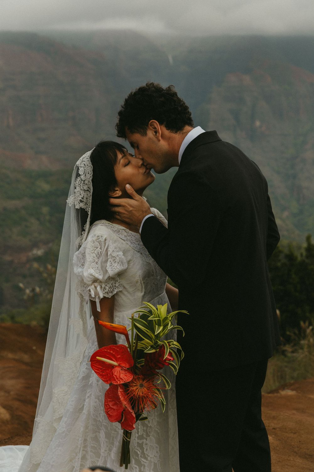 A romantic wedding couple shares a tender kiss on a scenic mountain overlook at sunset.