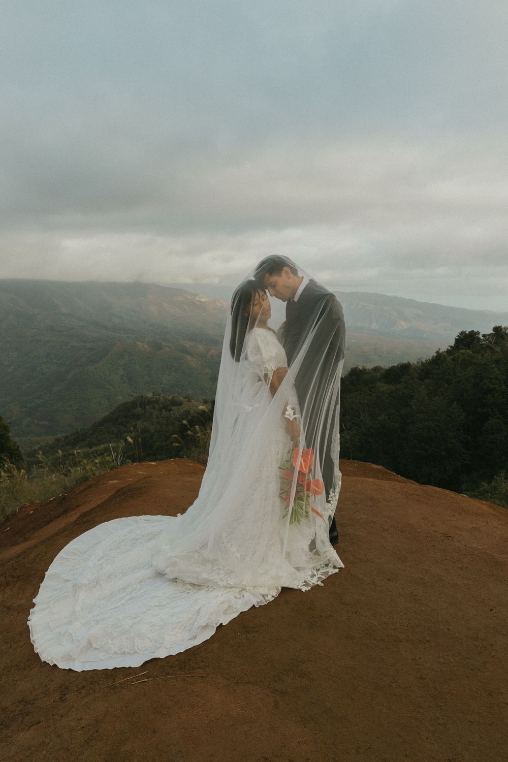 A couple in wedding attire embraces on a hilltop overlooking a misty mountain landscape at sunset.