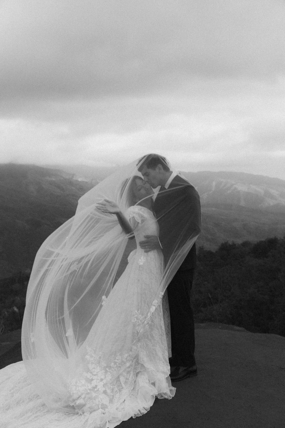 A romantic black and white wedding photo sequence showing a couple embracing as the bride's veil flows in the wind against mountains.