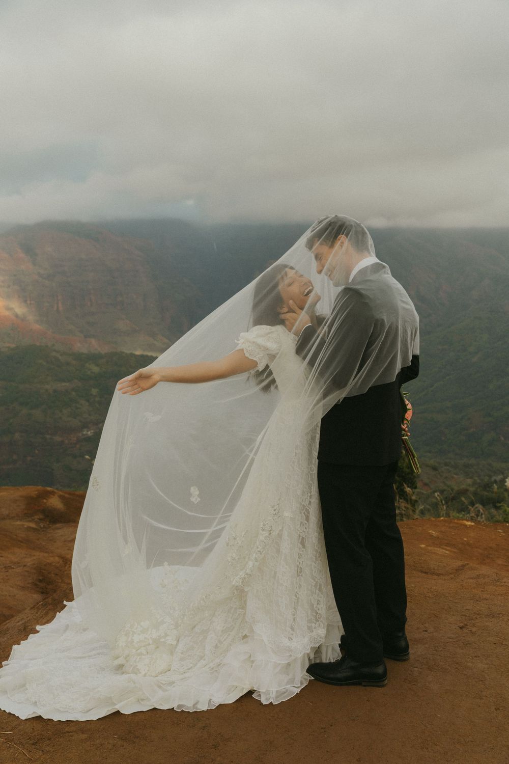 A couple in wedding attire shares a romantic moment on a mountain overlook with dramatic canyon views in the background.