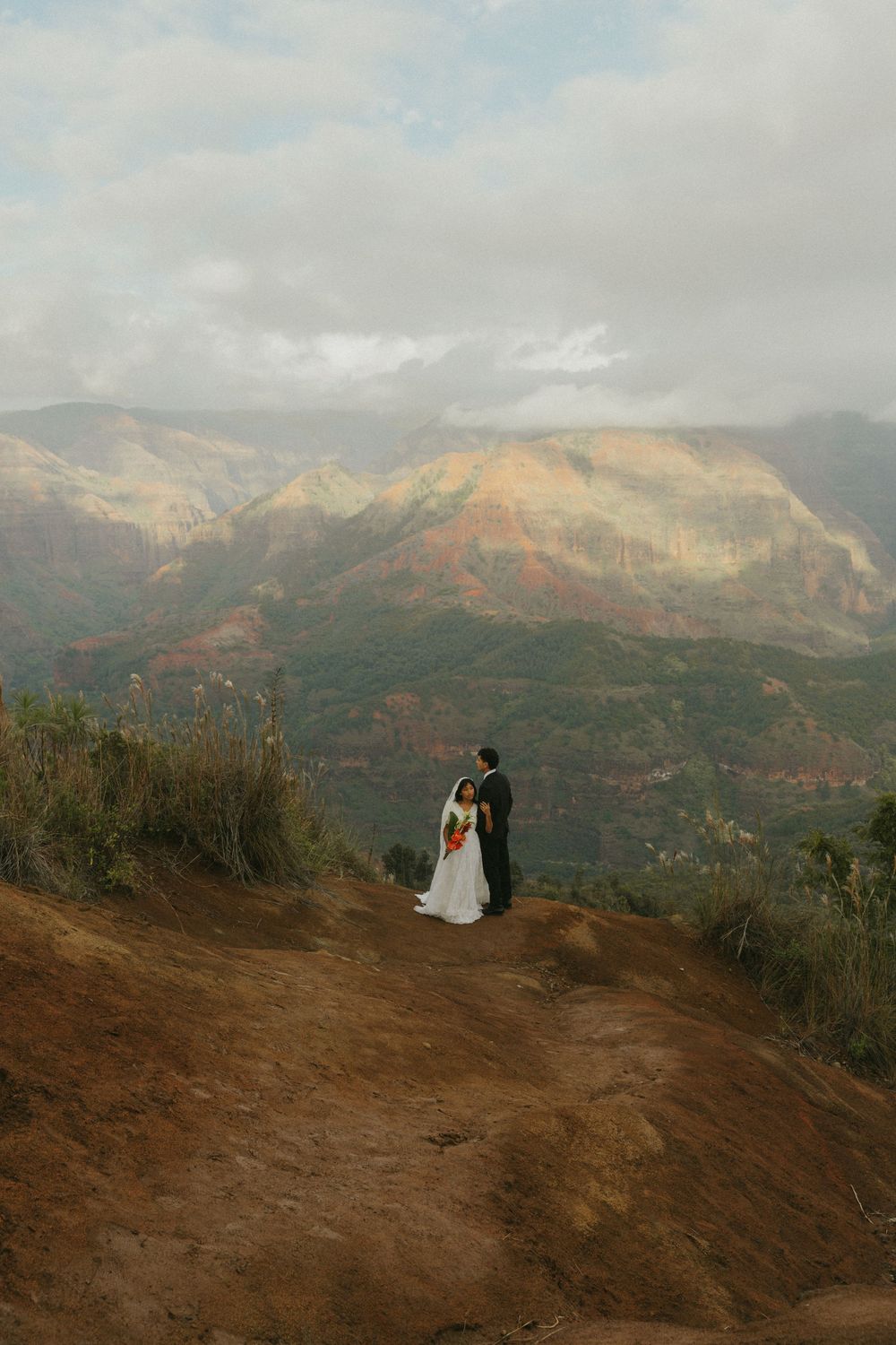A couple stands on a scenic mountain overlook with dramatic golden-hued peaks and cloudy skies in the background.
