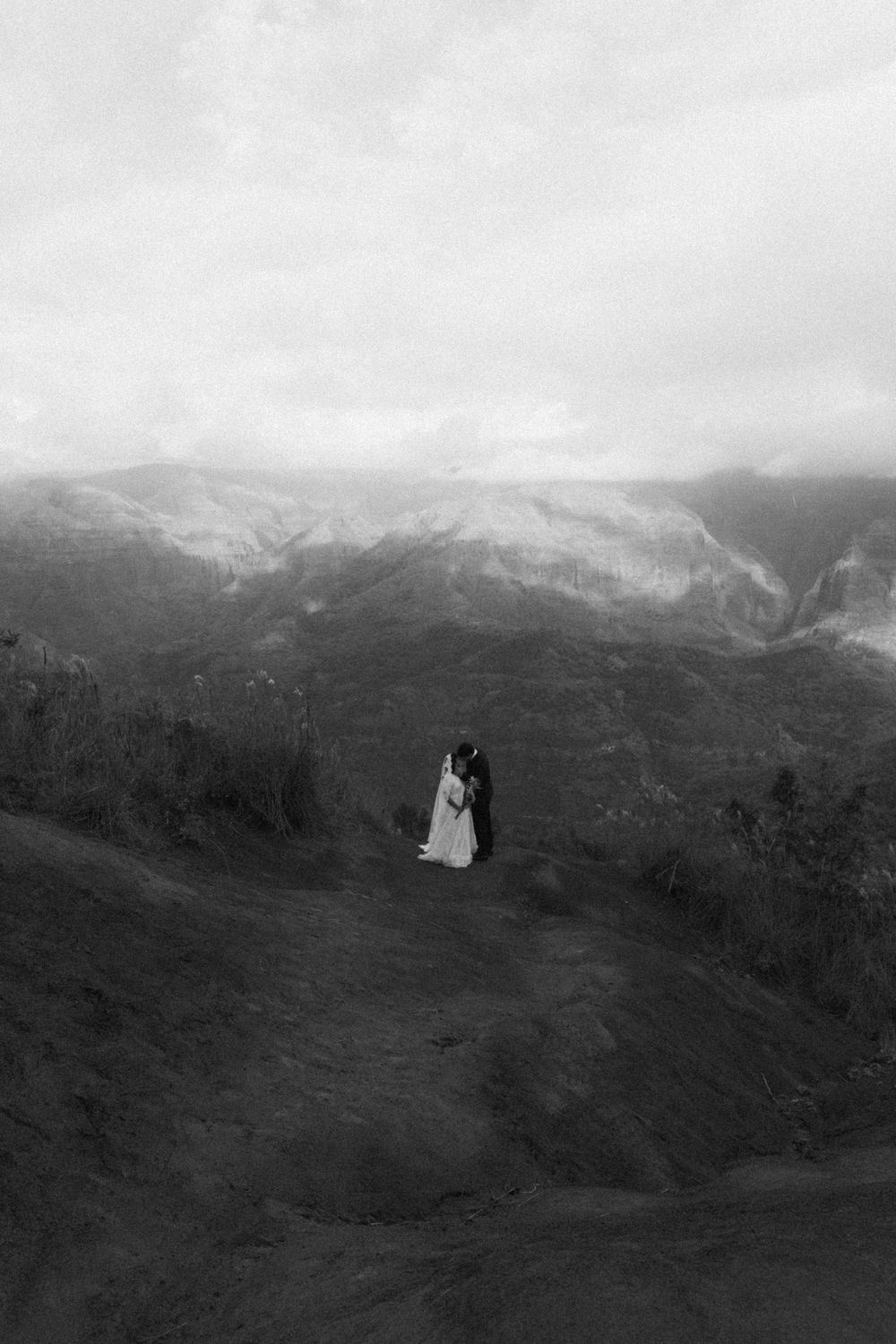 Dramatic black and white panoramic view of mountainous terrain with a distant wedding couple silhouetted.