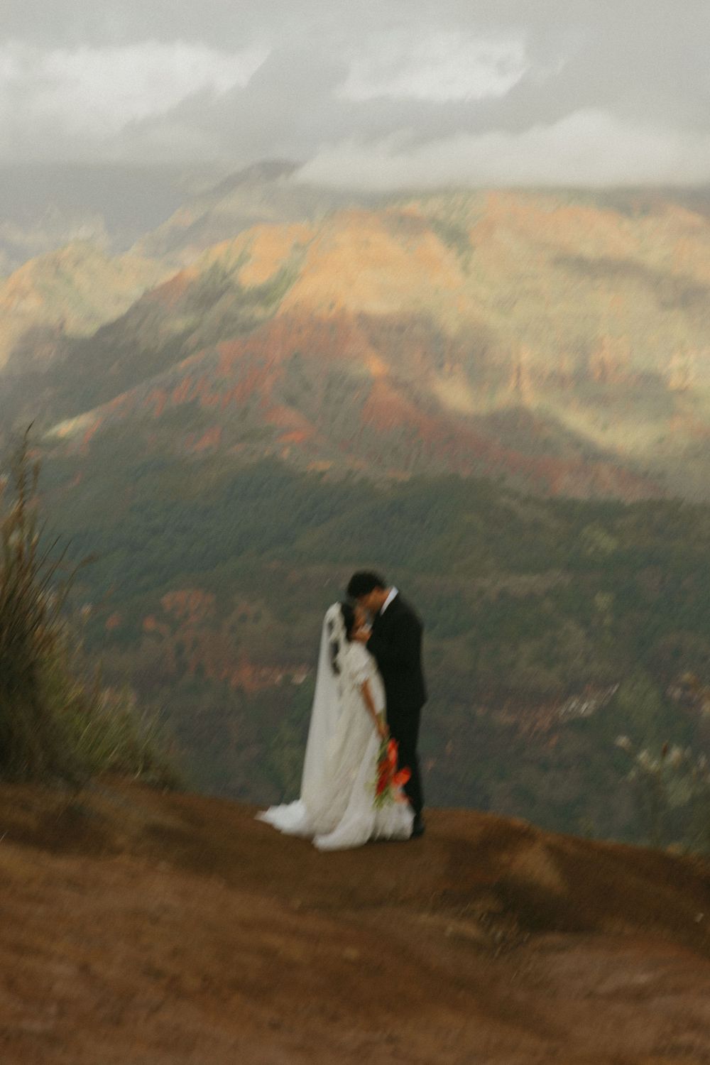 A couple in wedding attire stands on a mountain overlook with dramatic fall-colored peaks in the background at sunset.