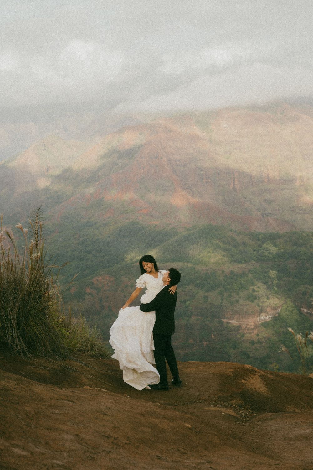 A couple in wedding attire sits on a mountainside overlooking a dramatic valley landscape at sunset.