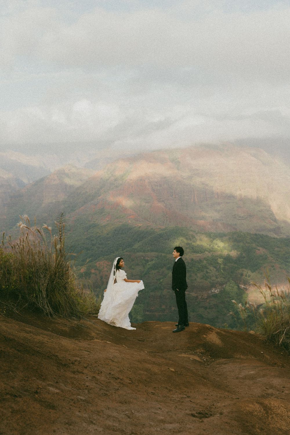 A couple in wedding attire poses on a scenic mountain ridge with dramatic cloudy skies in the background.