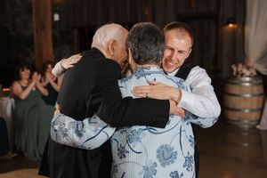 Three people share an emotional group hug at a rustic venue with wooden barrels visible in the background.