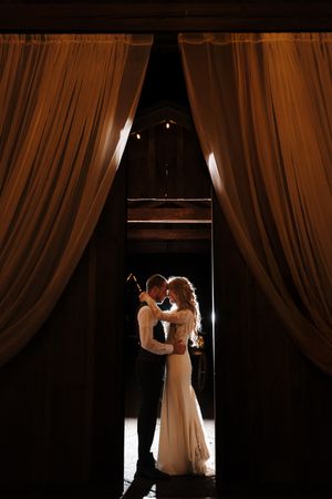 Romantic silhouettes of a couple dancing in elegant wedding attire against dramatic backlighting and flowing curtains.