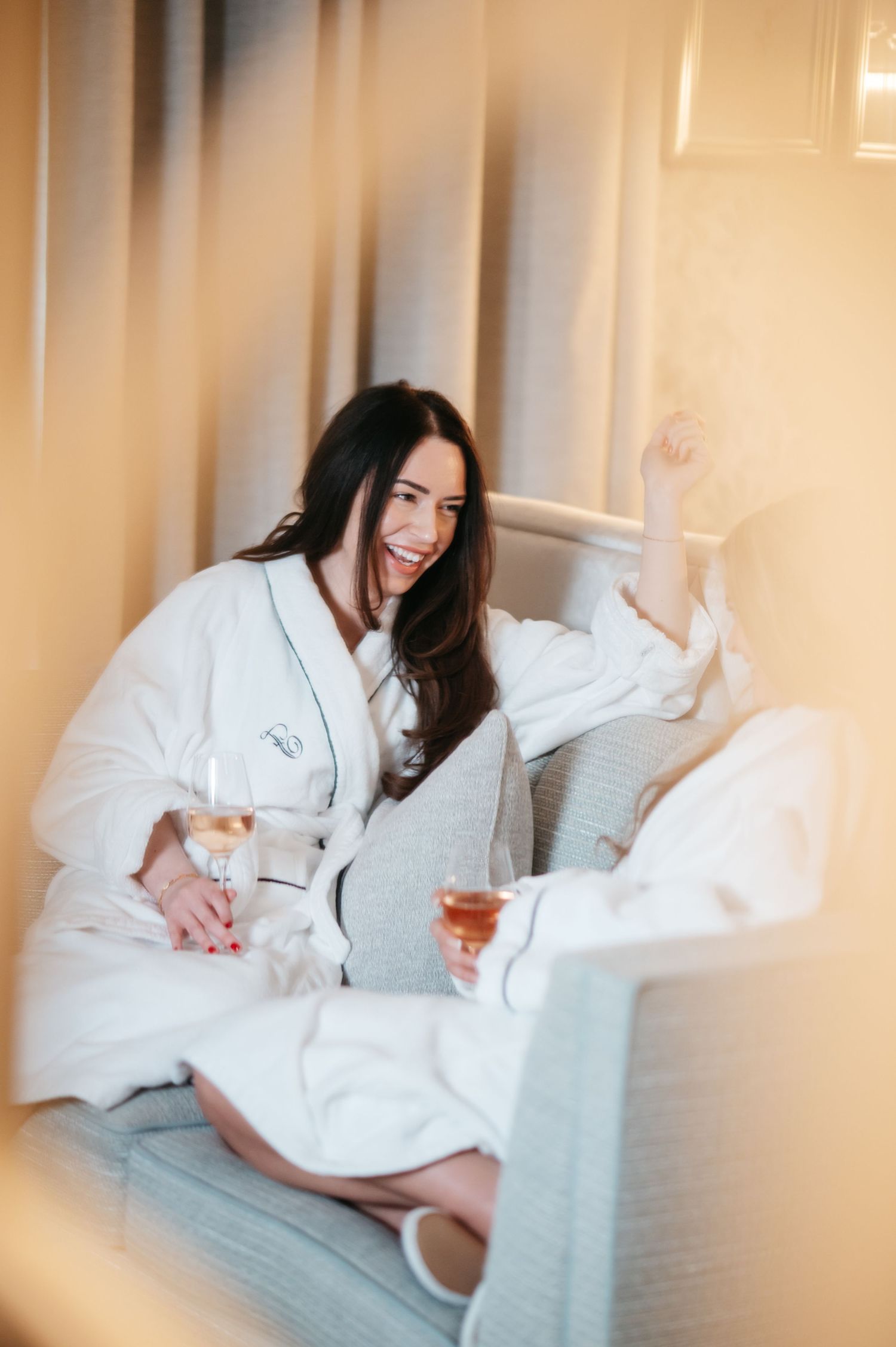 Smiling woman in white robe holding wine glasses, relaxing on a couch in a luxurious hotel room.