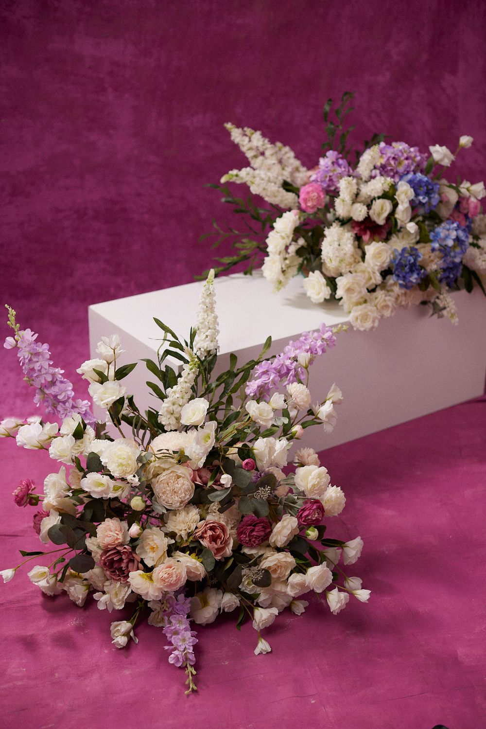 Two elegant floral arrangements with white, pink, and purple blooms displayed on a white pedestal against a magenta background.