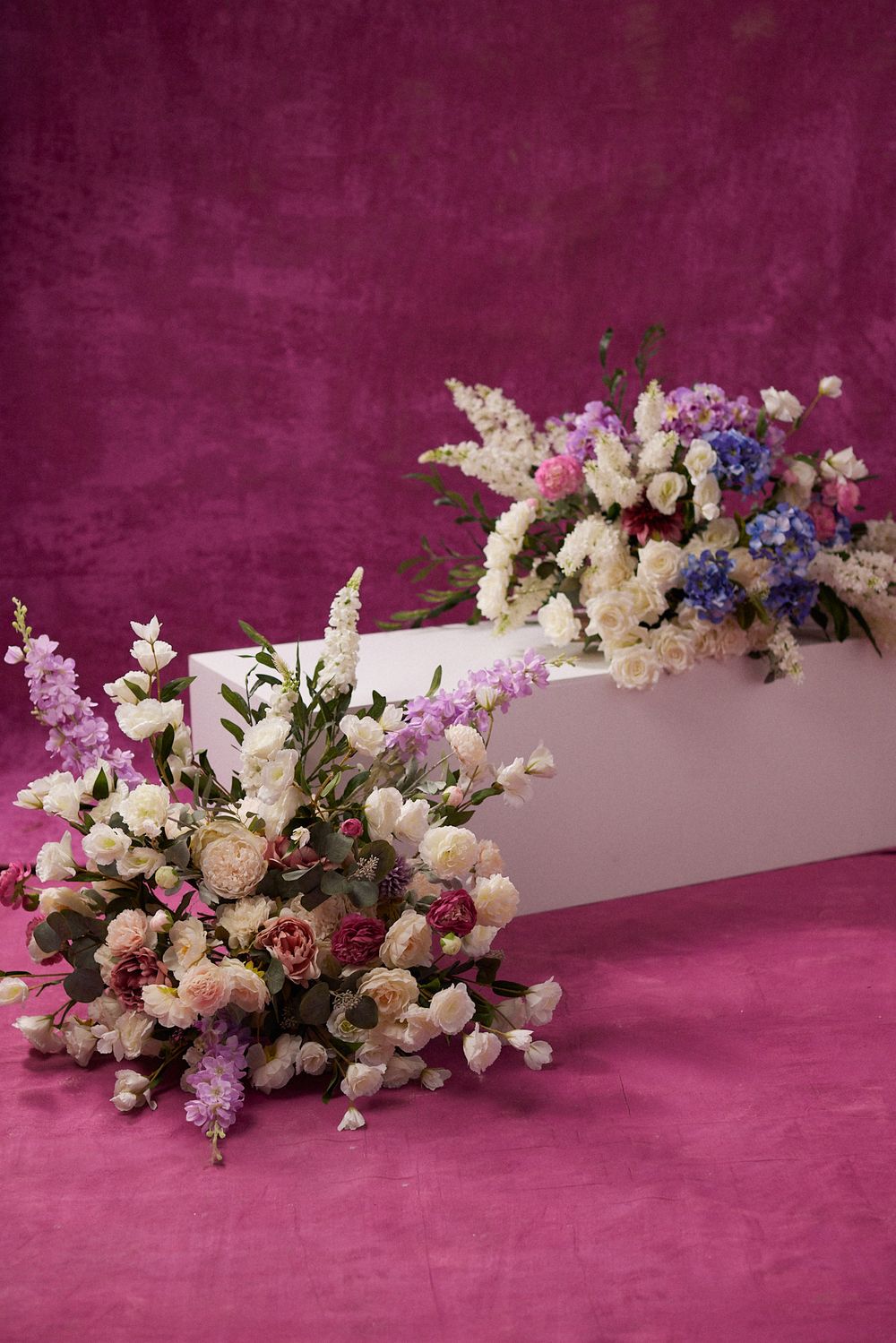 Two elegant floral arrangements with white, pink, and blue flowers displayed on a pink velvet background with a white pedestal.
