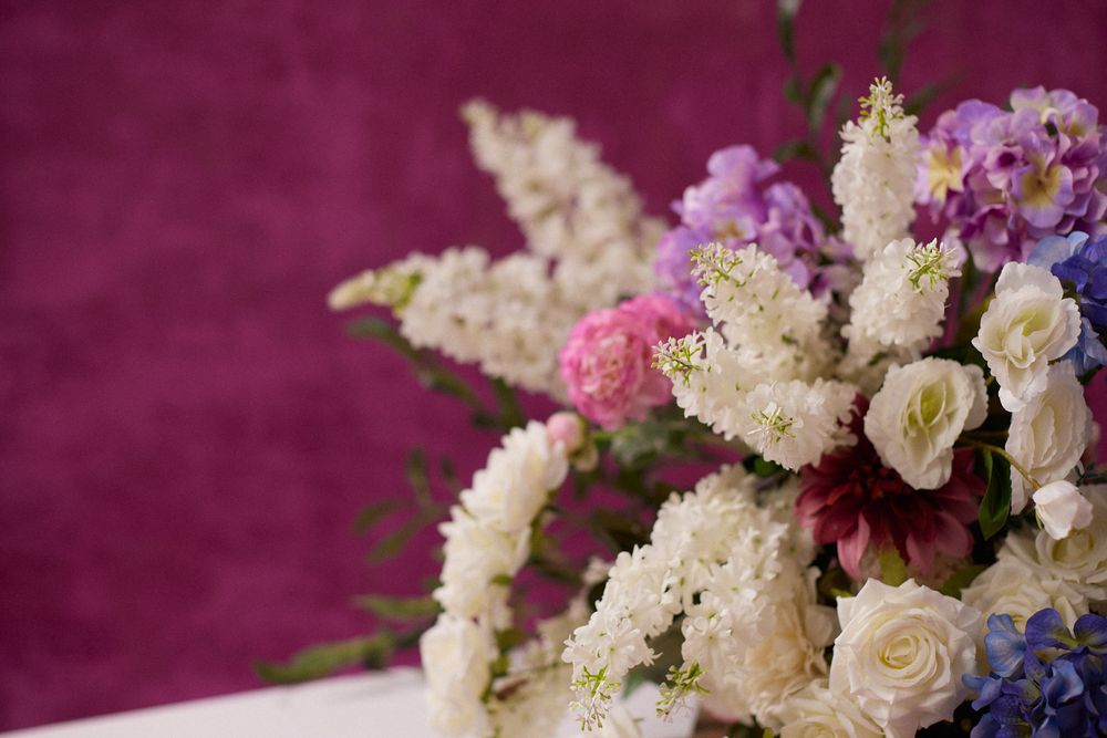 A lush bouquet of white roses, pink carnations, purple lilacs, and blue hydrangeas against a rich magenta background.
