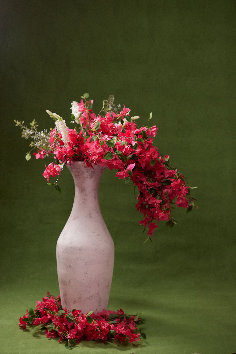 Pink vase with vibrant red bougainvillea flowers cascading over the sides on a green background.