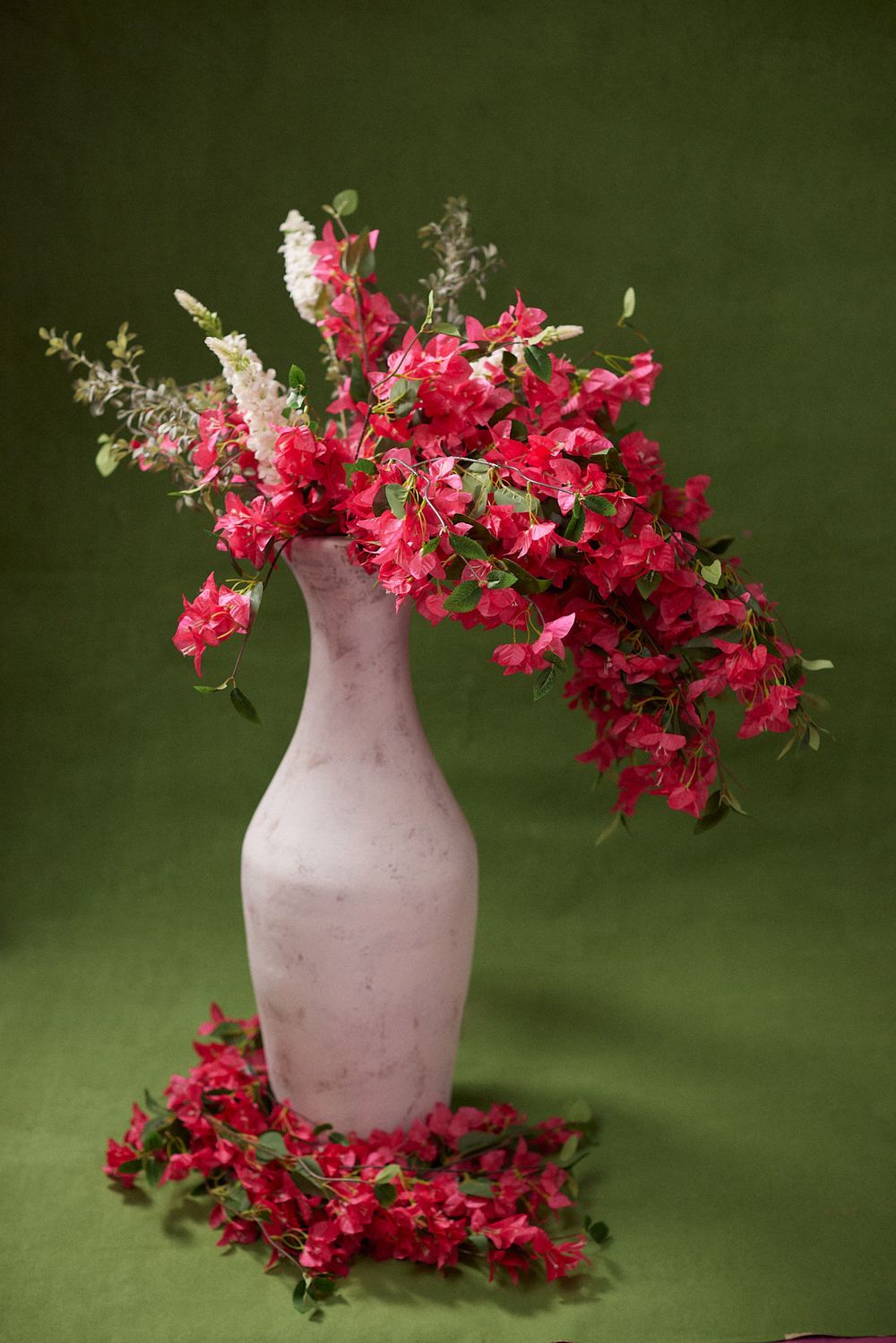 Pink vase with vibrant red bougainvillea flowers on green background, petals scattered around base.