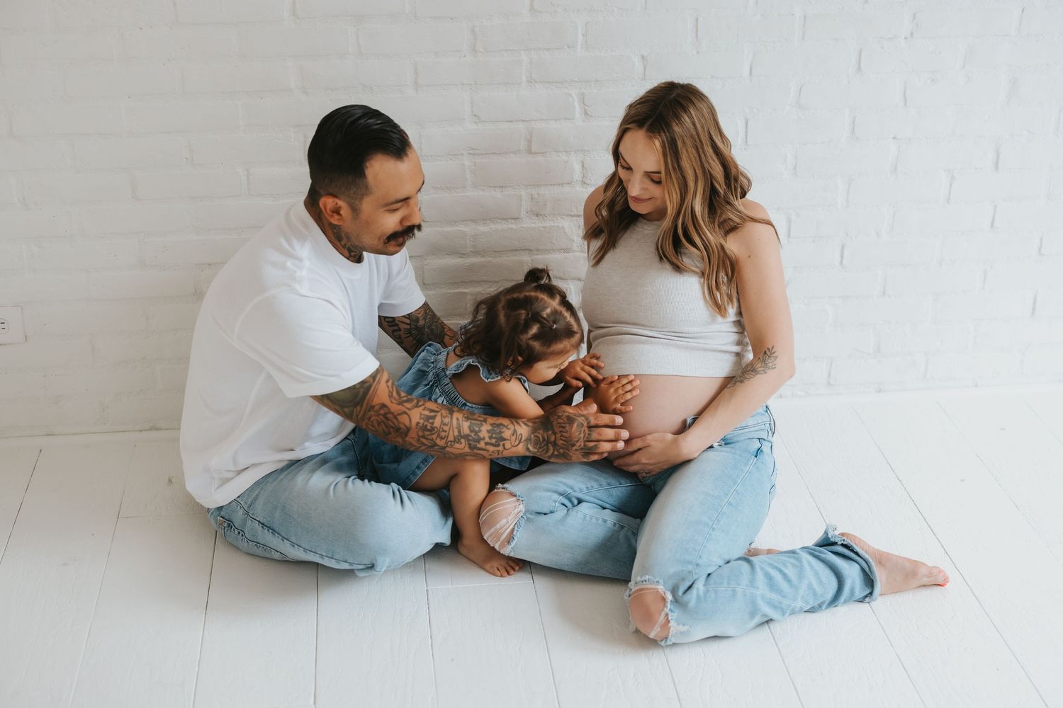 A couple holds and plays with a baby against a light gray wall in a series of candid lifestyle photos.