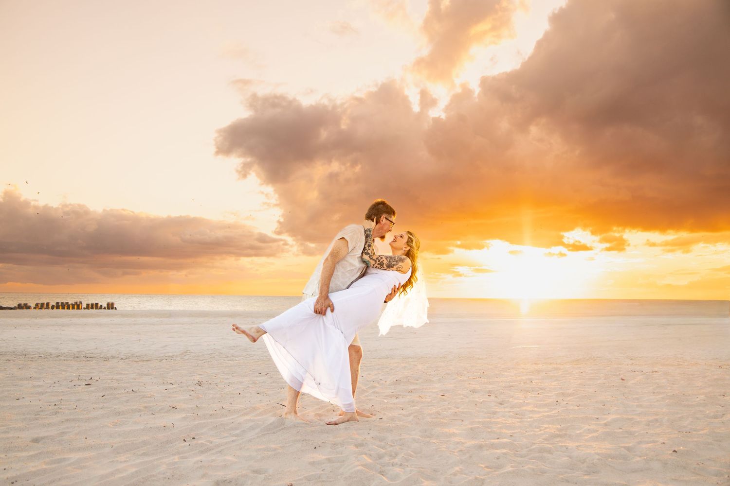 Couple sharing a romantic beach sunset embrace, groom lifting bride in white dress on sandy shore with golden sky. on Bradenton Beach