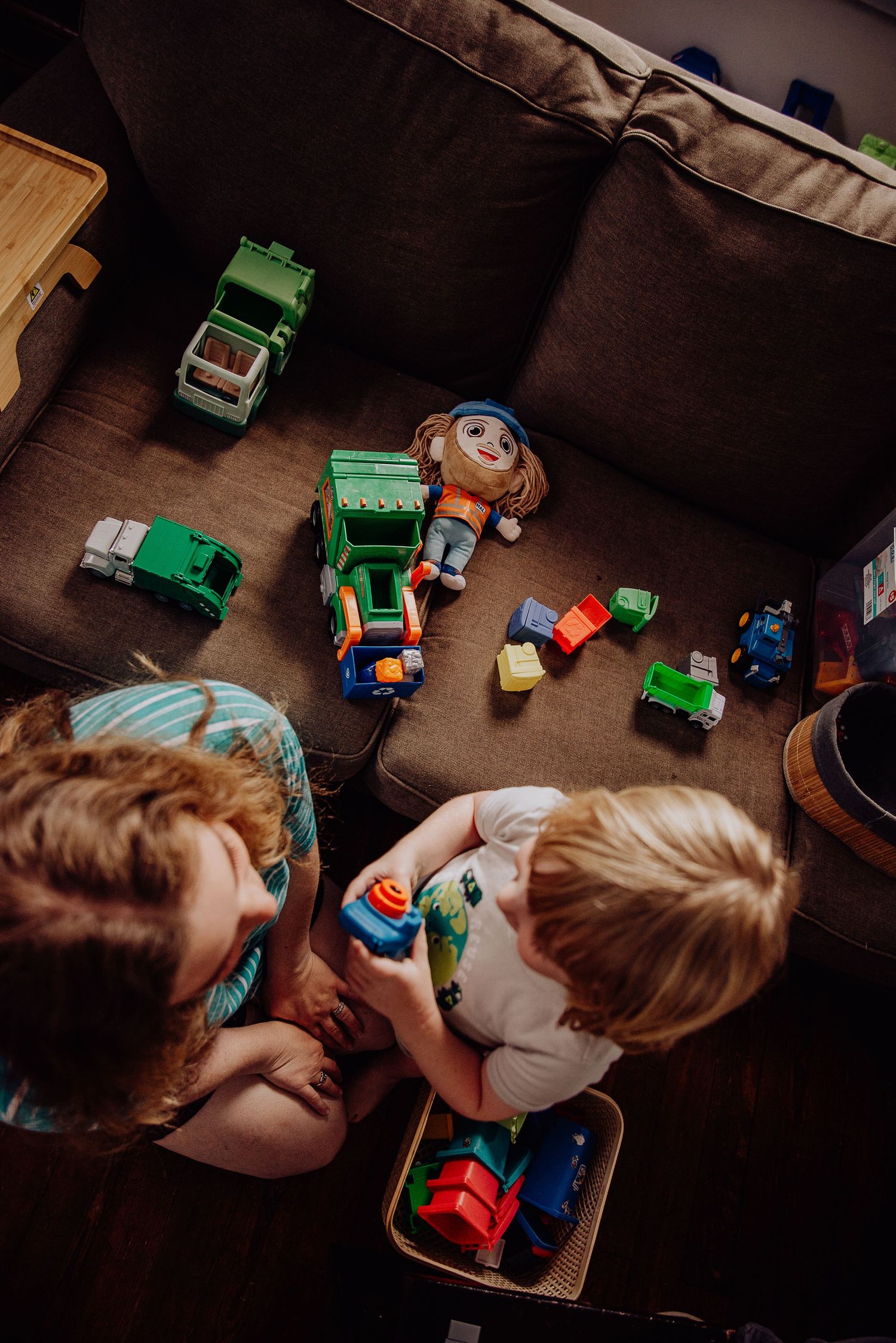 Two children playing with colorful toy vehicles and trucks on a brown couch in a dimly lit room.