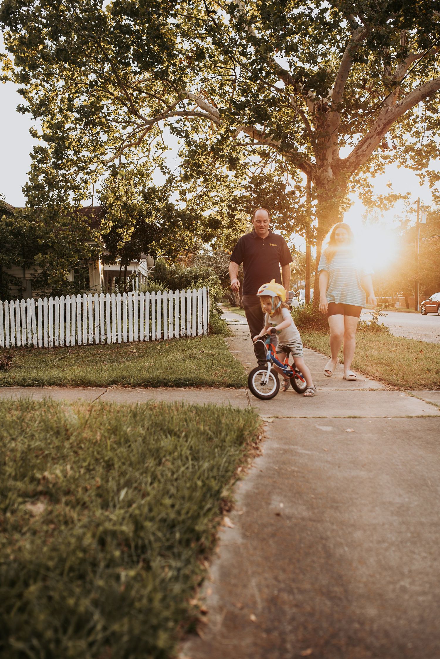 A child learns to ride a bike on a sunset-lit sidewalk path while being guided by an adult on a warm evening.
