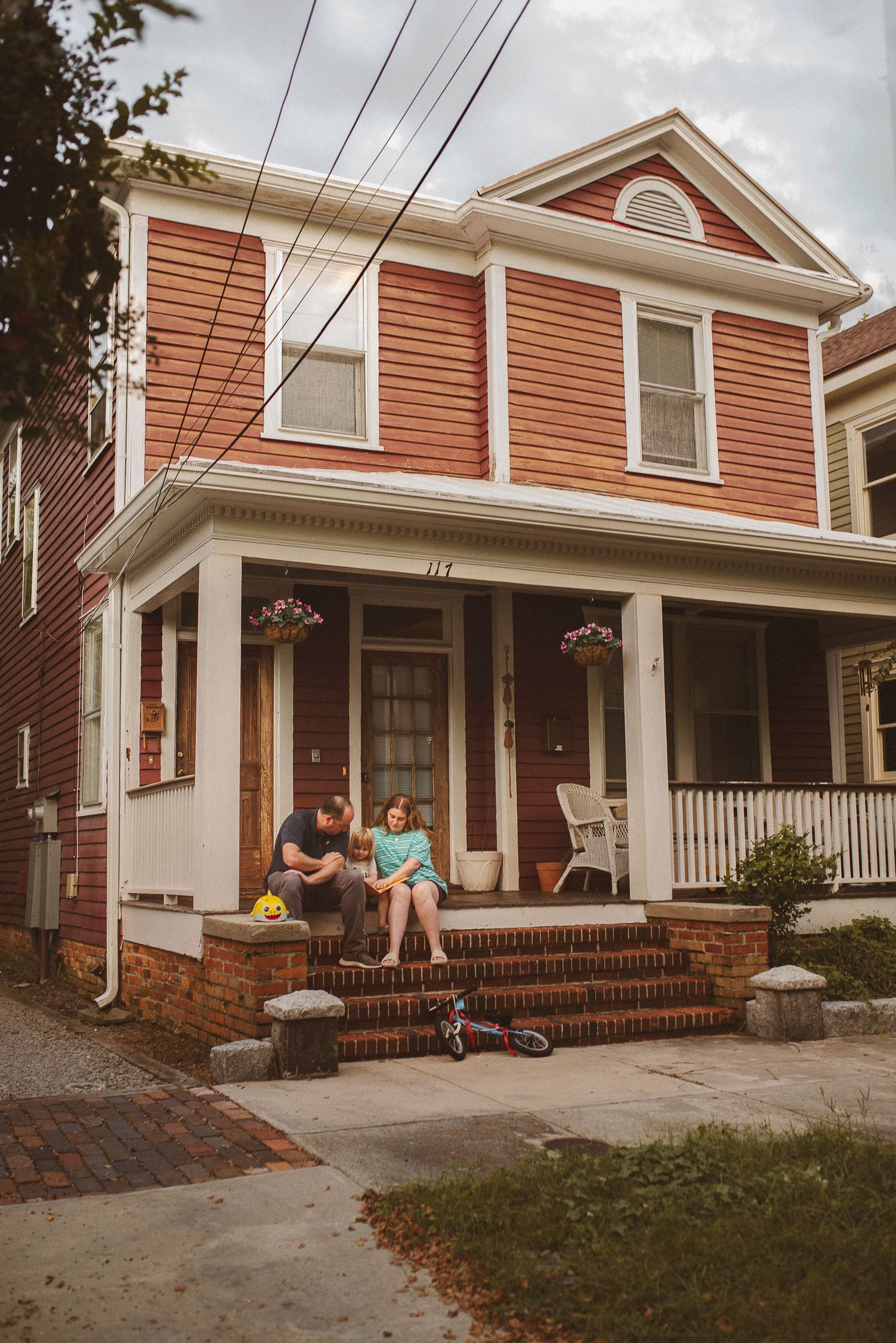 People relaxing on the front porch steps of a traditional red-sided house with white trim on a cloudy day.