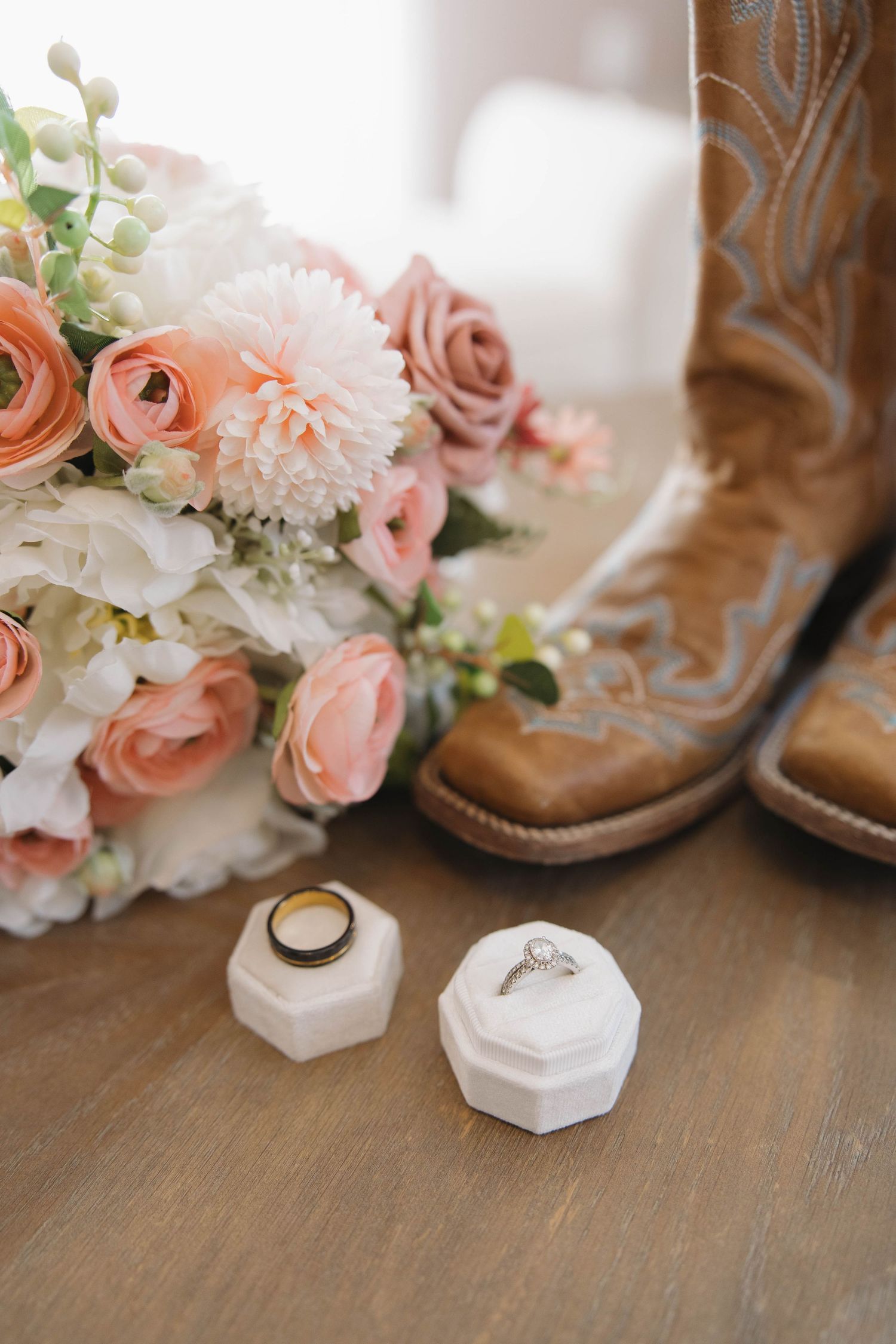 Wedding details with brown cowboy boots, pink and white flowers, and ring boxes on wooden surface.