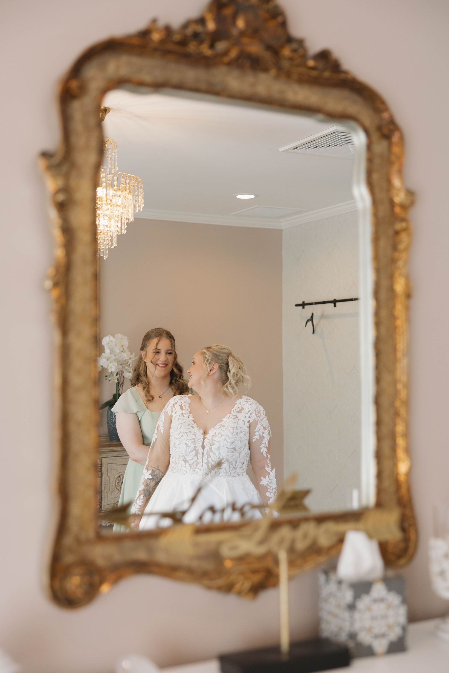 Ornate gold-framed mirror reflects a bridal preparation scene in an elegant white room.