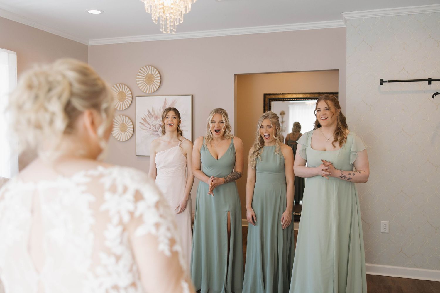 Bridesmaids in sage green dresses stand together in an elegant room with a crystal chandelier.