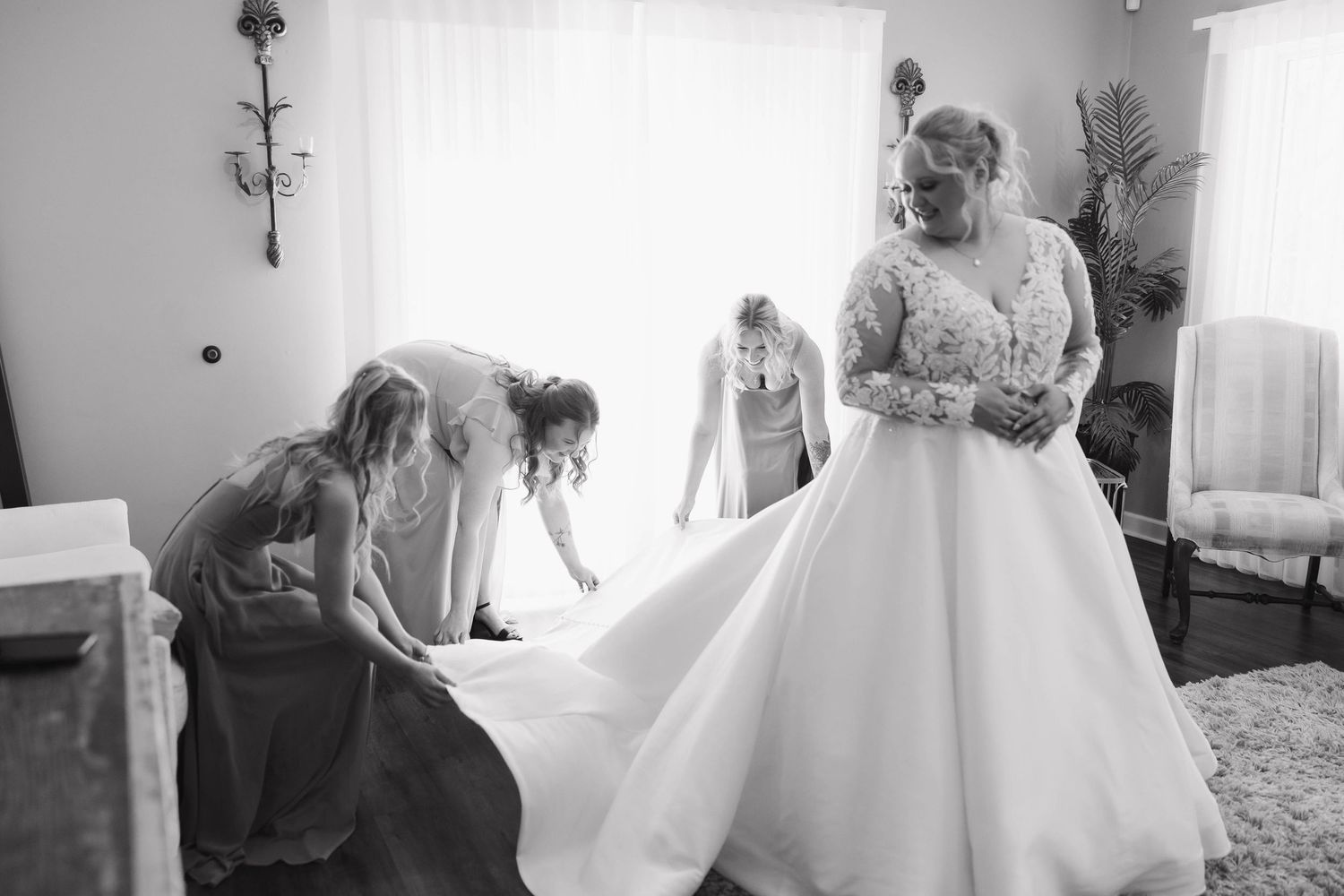 A bride in a white wedding gown gets ready with help from others in a bright sunlit room.