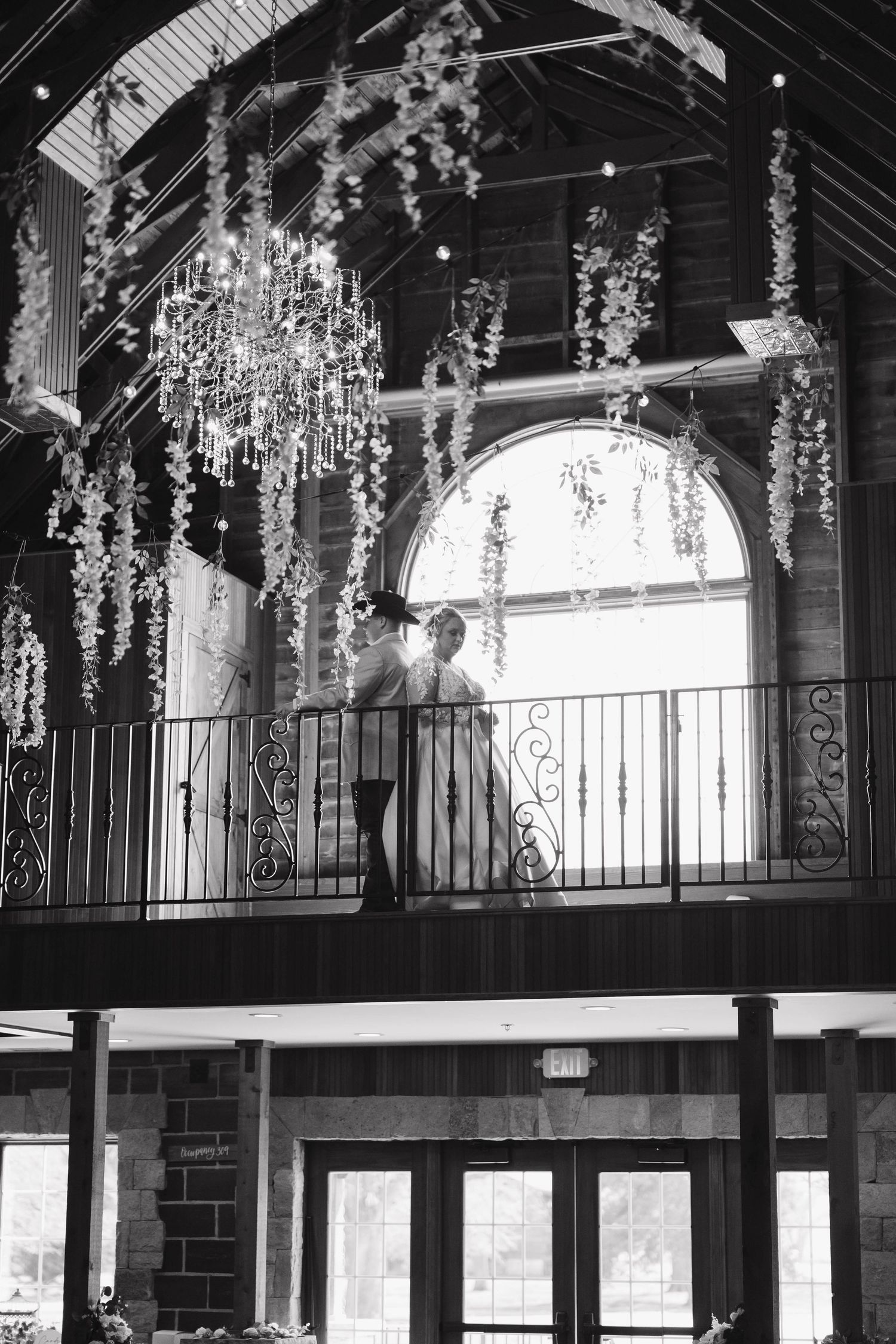 Black and white photo of a rustic wedding venue interior with hanging chandeliers and decorative floral garlands.
