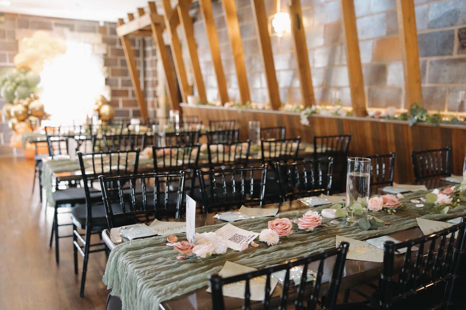 Long banquet tables with black chairs set up in a rustic brick-walled reception venue with natural lighting.