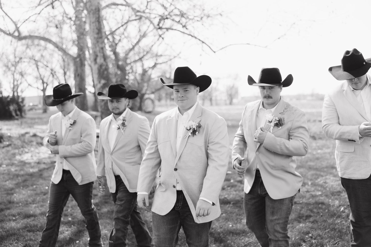 Groomsmen in white jackets and cowboy hats run playfully through a field during a rustic outdoor wedding.