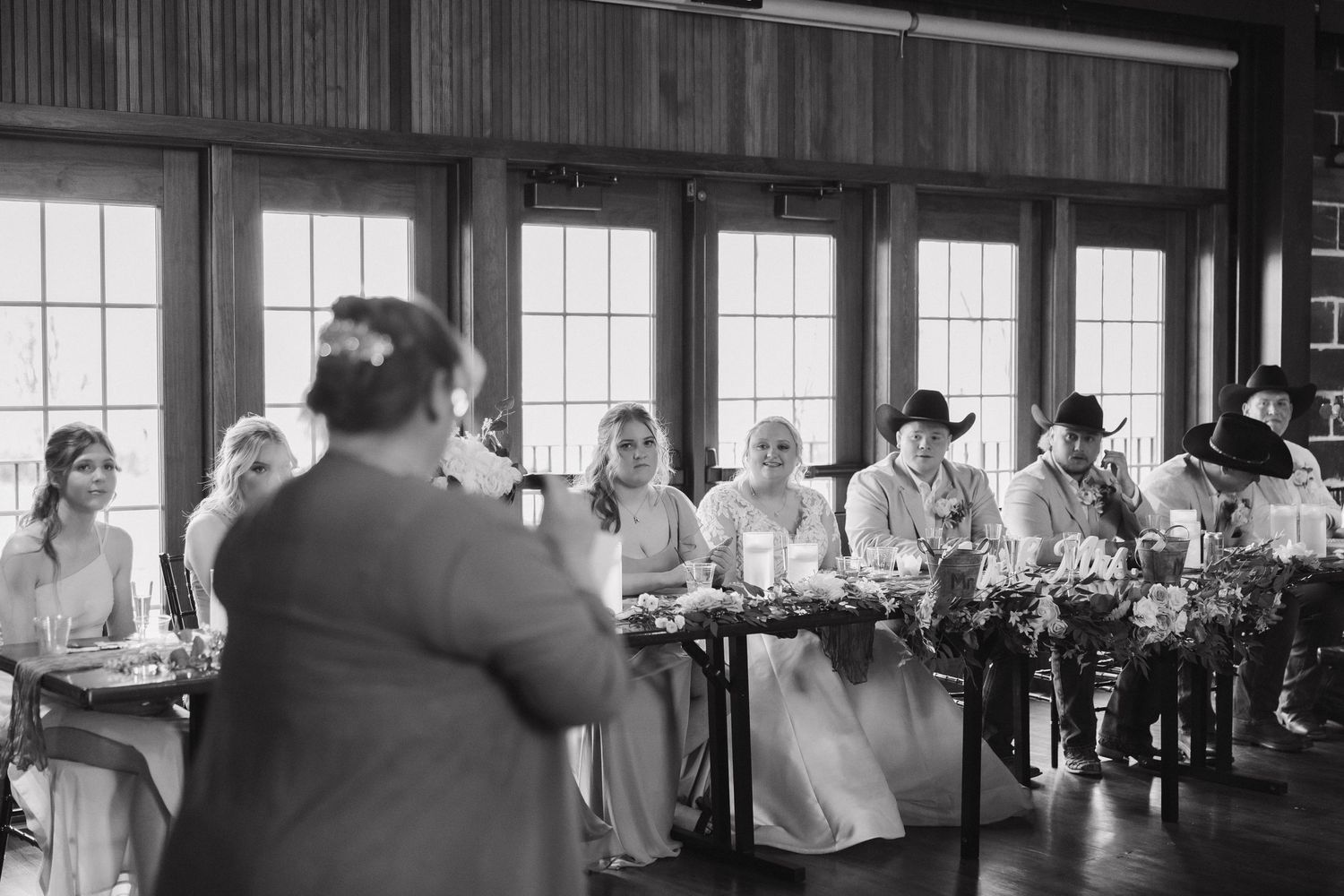 Black and white photo of wedding reception with guests seated at long tables in rustic venue.