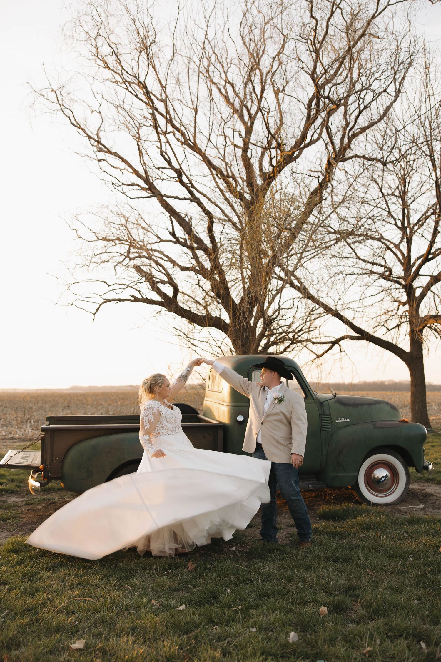 A romantic countryside photoshoot featuring a vintage green truck and flowing white dress in a rustic field at sunset.