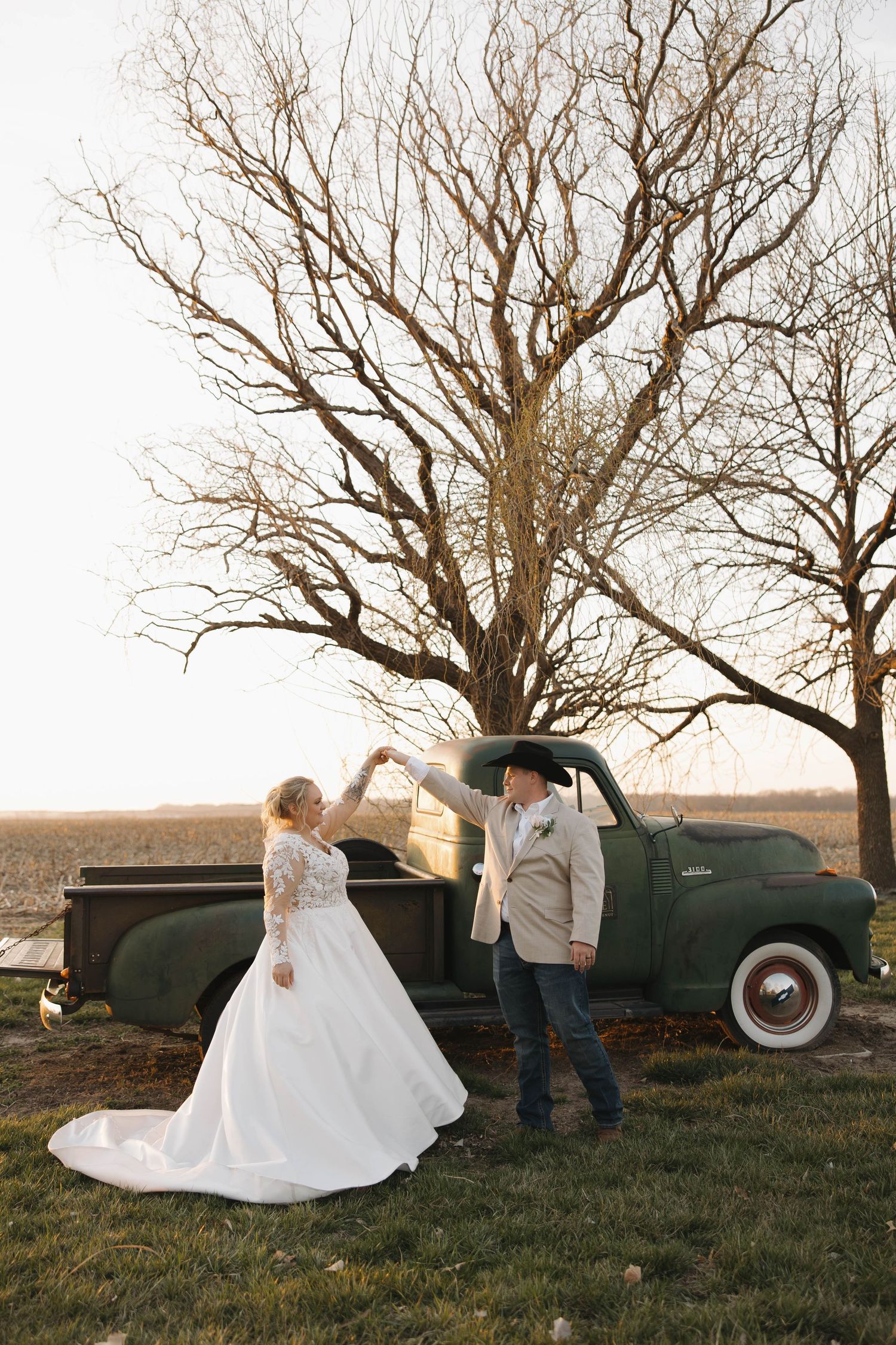 Wedding couple poses by vintage truck in rustic outdoor setting with bare trees.