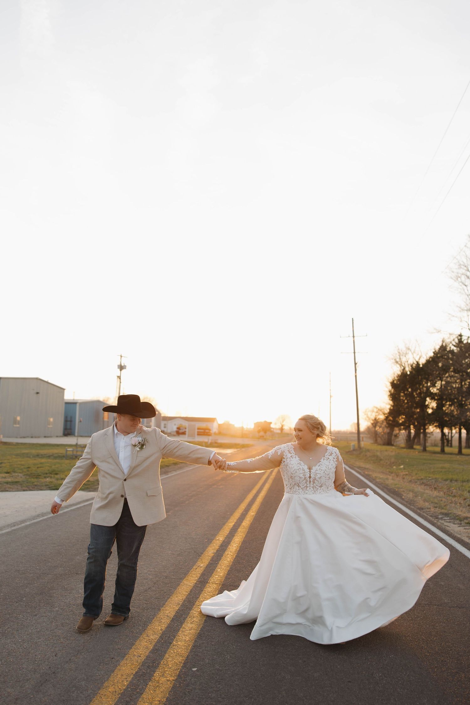 A couple dances and twirls on a country road at golden hour, with the bride's white dress flowing in the sunlight.