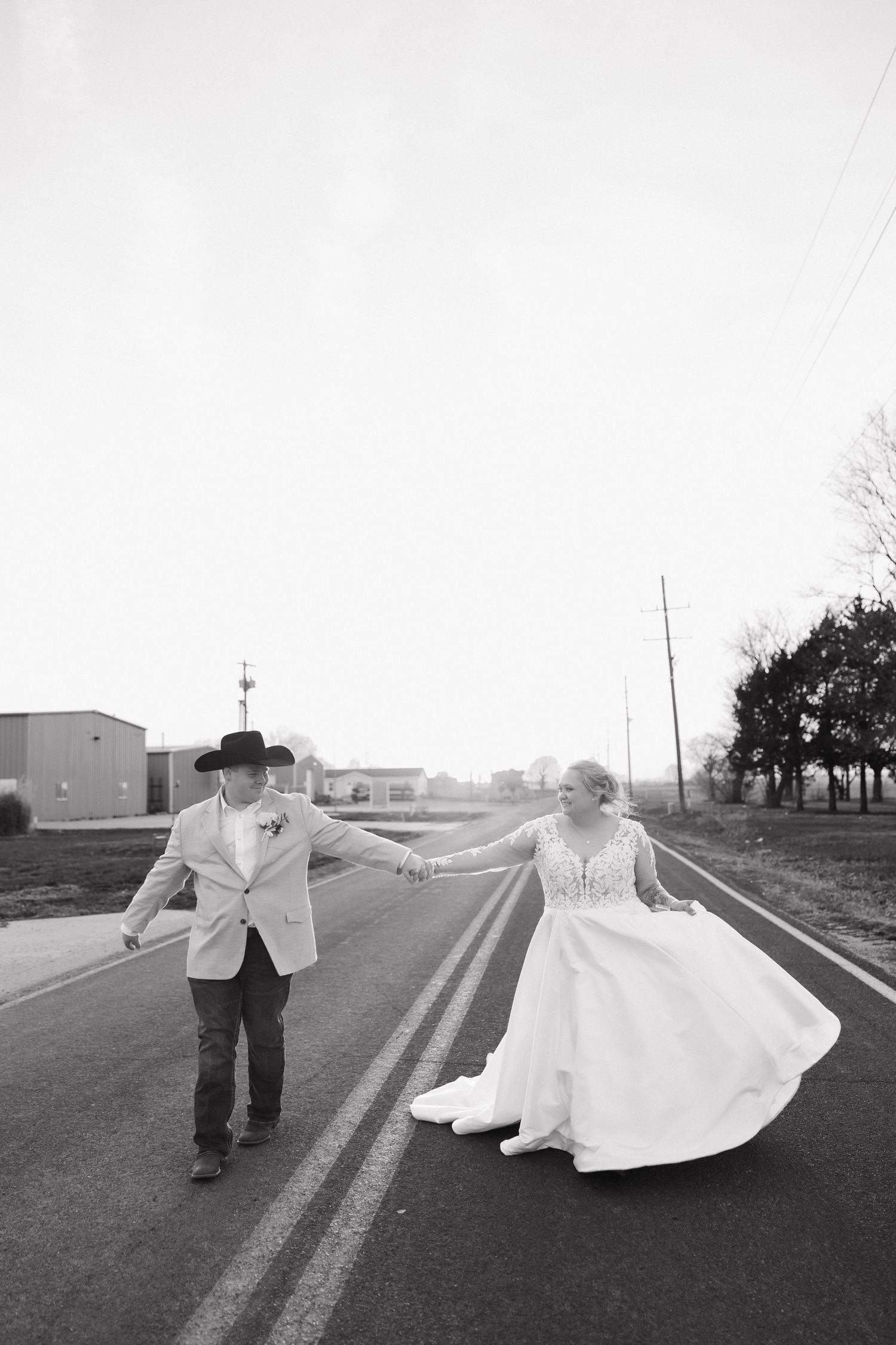 A joyful couple in wedding attire dances and twirls together on an empty rural road in black and white.