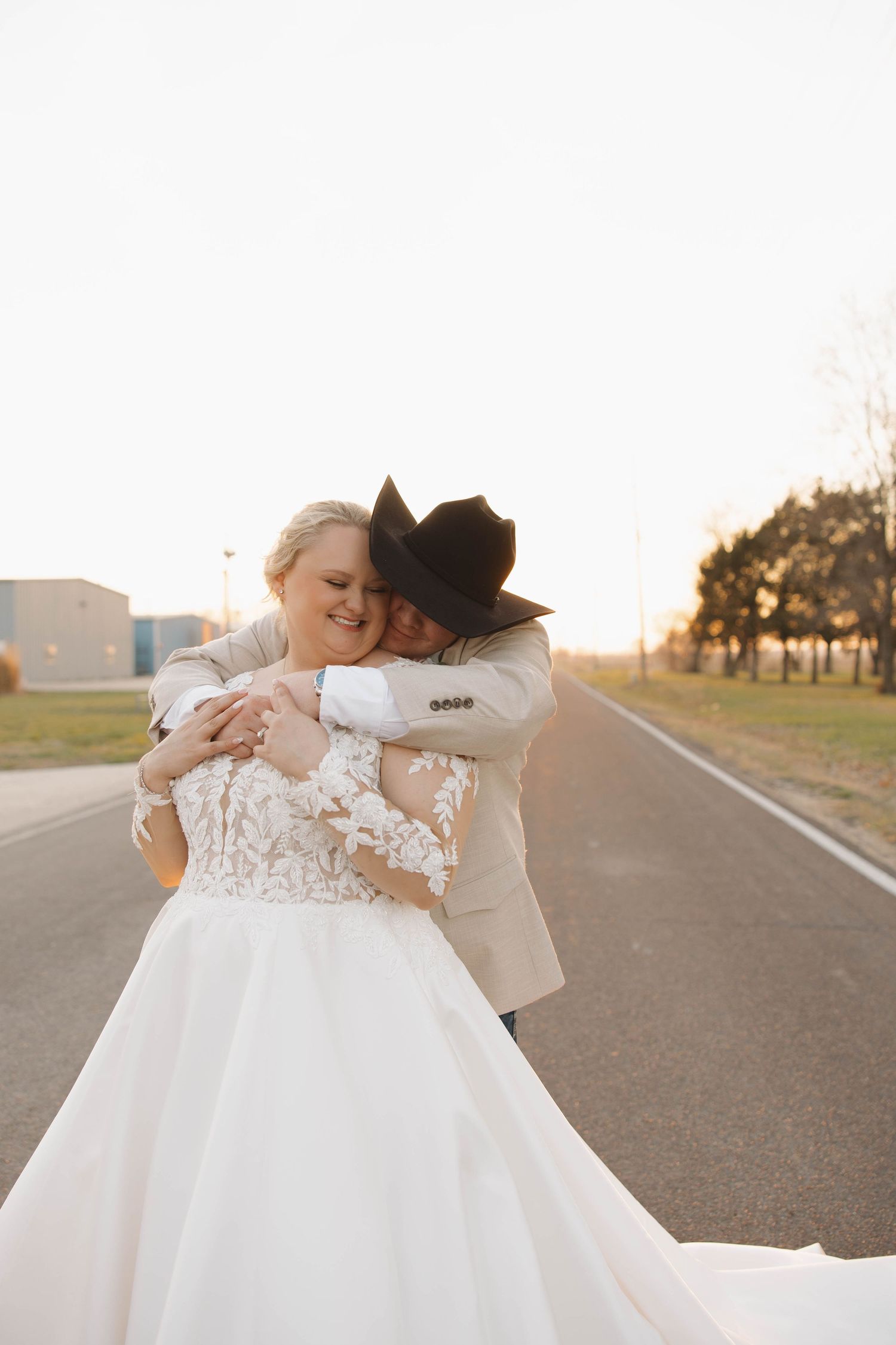 Couple embracing on empty road at sunset, bride in lace wedding gown and groom in cowboy hat.