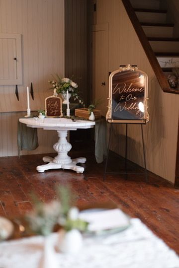 Rustic wedding venue entrance with white pedestal table, candles, florals, and illuminated welcome mirror sign.