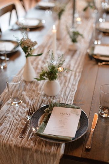 Rustic wedding table setting with dark charger plates, menu cards, white bud vases, and cheesecloth runner.