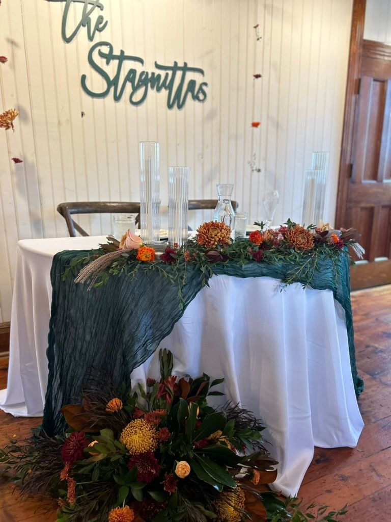 Wedding sweetheart table with teal cheesecloth runner, fall floral garland, and The Stagnittas wooden name sign.