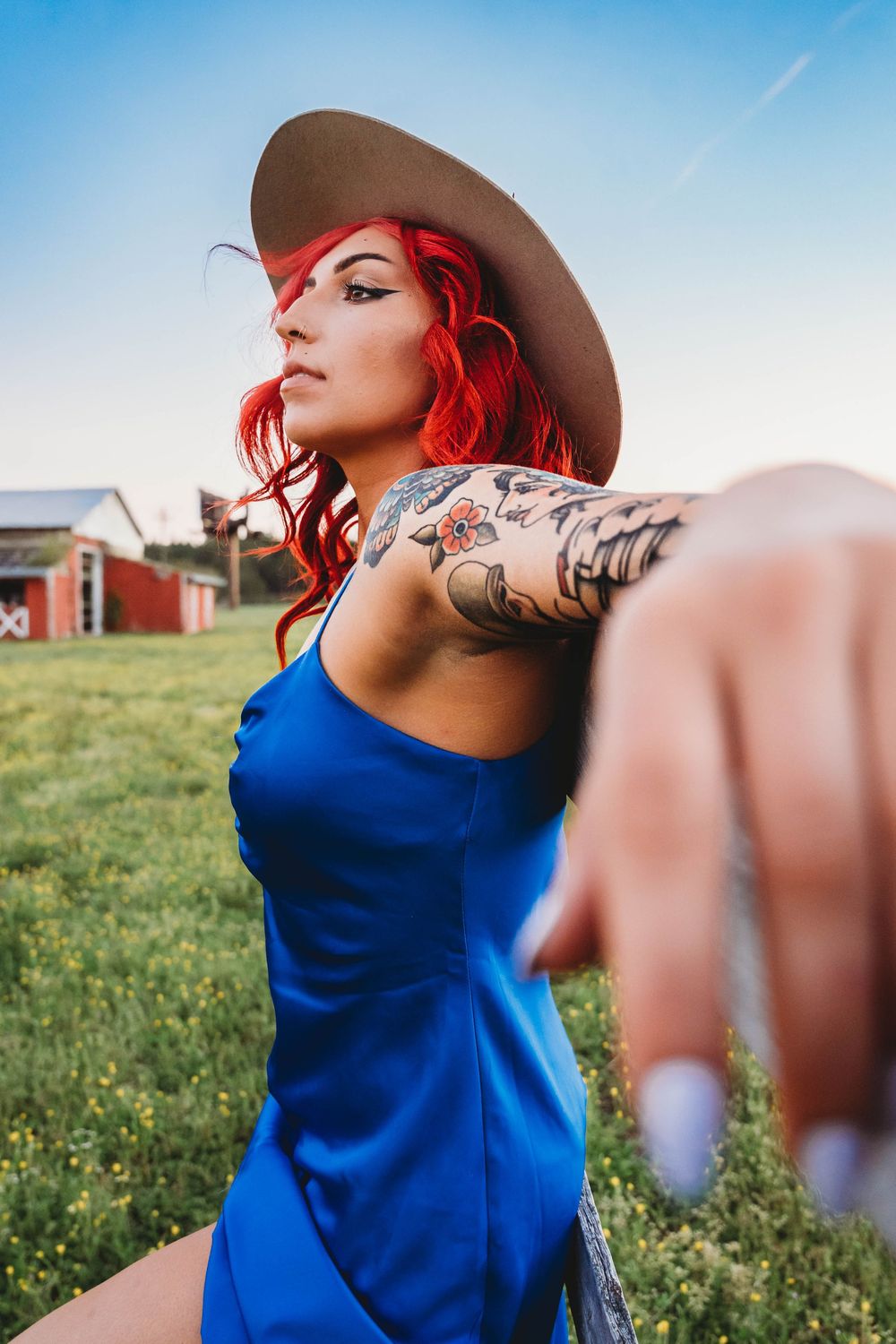 Red-haired woman in blue dress and brown hat with tattoos reaching toward camera in a rural farm setting.