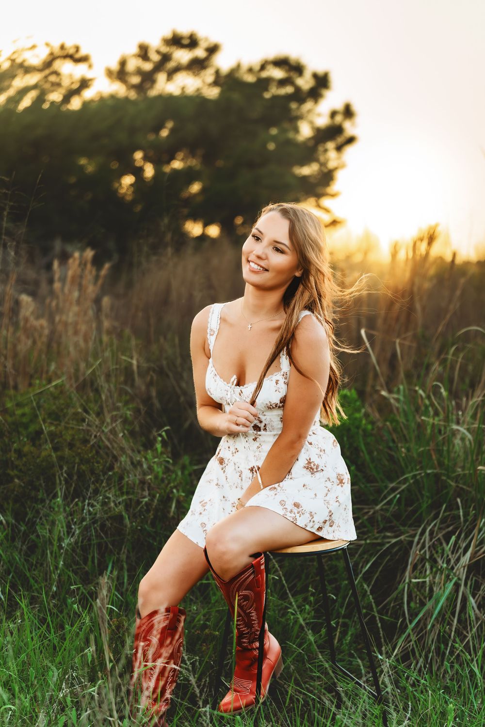 Young woman in floral dress and red cowboy boots sitting in a sunlit field at golden hour, smiling warmly.