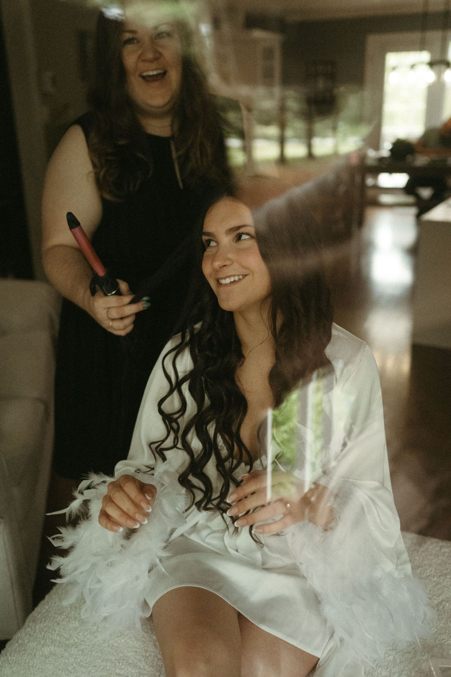 Bride smiling in white robe while hairstylist curls her long dark hair for wedding.