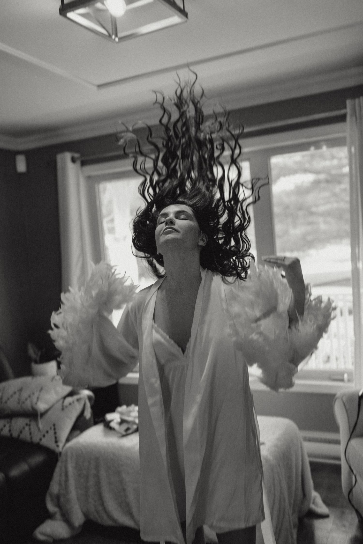 Black and white photo of bride in feathered robe tossing long curly hair dramatically indoors.