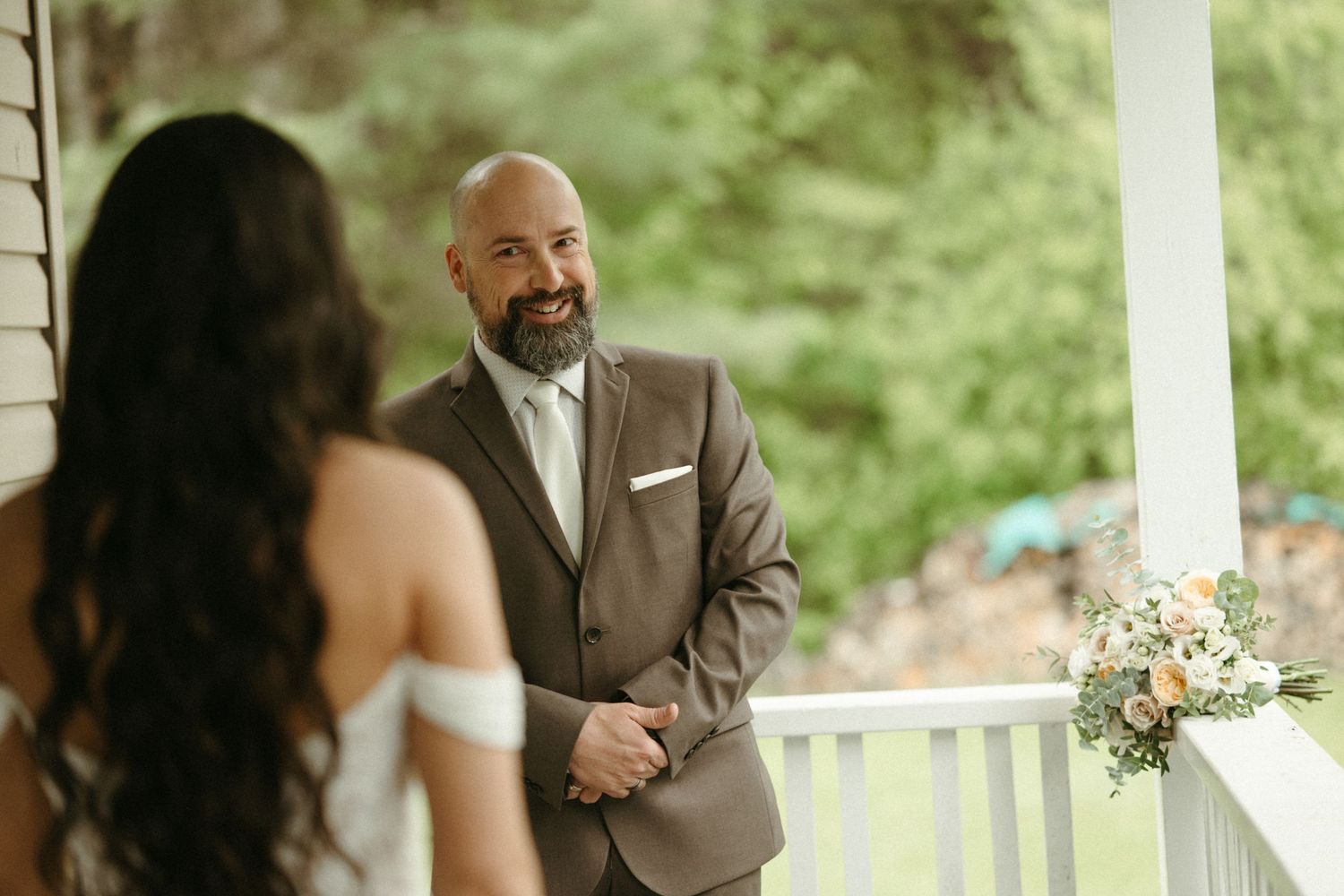 Groom in brown suit smiling at bride during outdoor wedding first look on white porch.