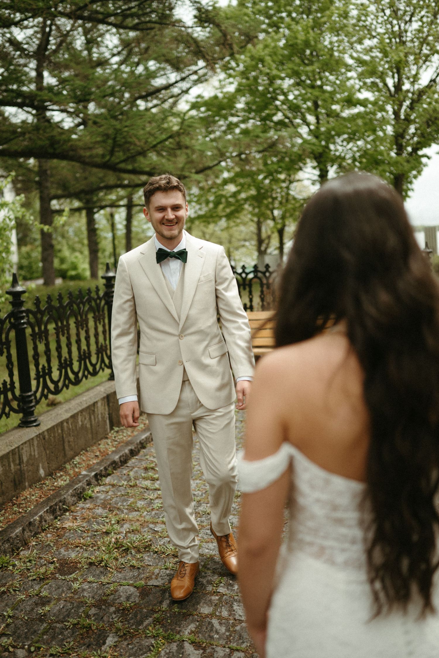 Smiling groom in beige suit with green bow tie walks toward bride during first look.